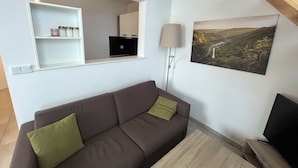 Ein Wohnzimmer mit einem braunen Sofa, einer kleinen weißen Regalanlage, einer Lampe und einem Landschaftsbild.