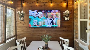 A wooden-paneled room with a TV displaying a sports game, a white table with chairs, and a hanging light fixture. - Lone Oak Cottage Fully restored cabin with 5 beds & walk in shower (Medicine Park)