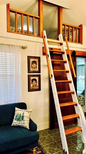 A loft-style room with a wooden staircase, a blue sofa with a 'WELCOME to our neck of the woods' pillow, and two framed pictures on the wall.