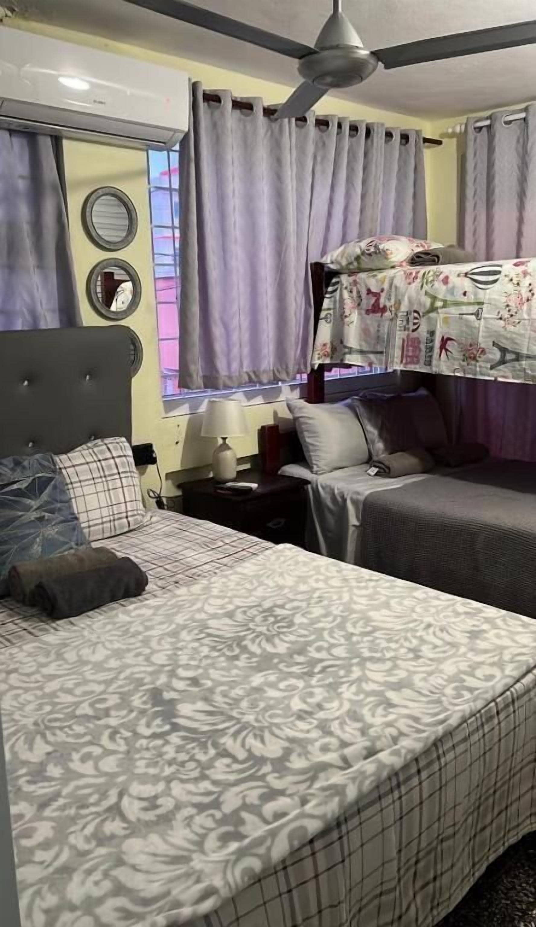 A hotel room with two beds, a ceiling fan, a bedside table with a lamp, and a window with curtains.