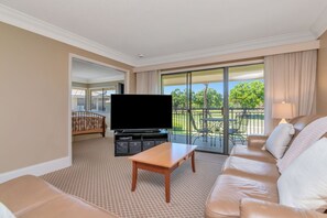 A hotel room with a large TV, a sofa, a coffee table, and a balcony with outdoor furniture.