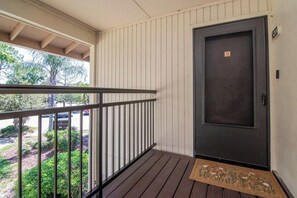 Eine Holzterrasse mit Metallgeländer, eine dunkle Tür mit Fußmatte und Blick auf Grünflächen und geparkte Autos.