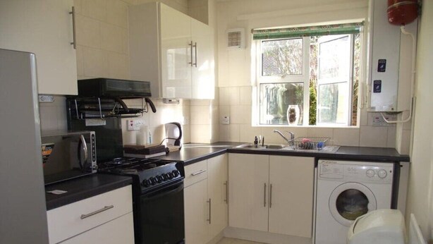 A compact kitchen with a stove, oven, washing machine, and a window with a view of greenery. - A good flat close to university! (Manchester)