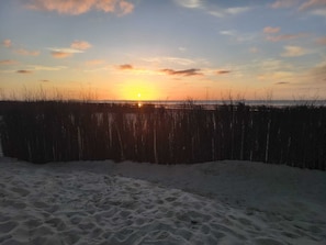 Ein Strand mit Sand und einem Zaun im Vordergrund, sowie ein Sonnenuntergang im Hintergrund.