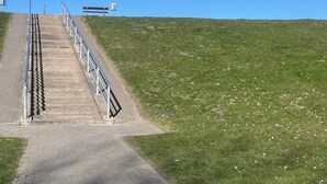 Ein gepflasterter Weg führt auf einen grasbewachsenen Hügel, der mit Metallgeländern und einer Bank an der Spitze ausgestattet ist.