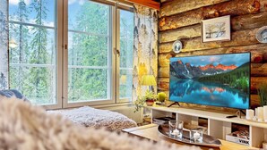 Interior de una cabaña con un ventanal grande, paredes de madera, un televisor mostrando un paisaje montañoso y una zona de estar acogedora.