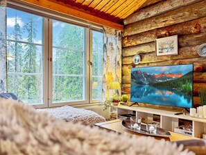Ein Urlaubsdomizil mit großem Fenster, Holzwänden, einem Fernseher, der eine Berglandschaft zeigt, und einer gemütlichen Sitzecke.