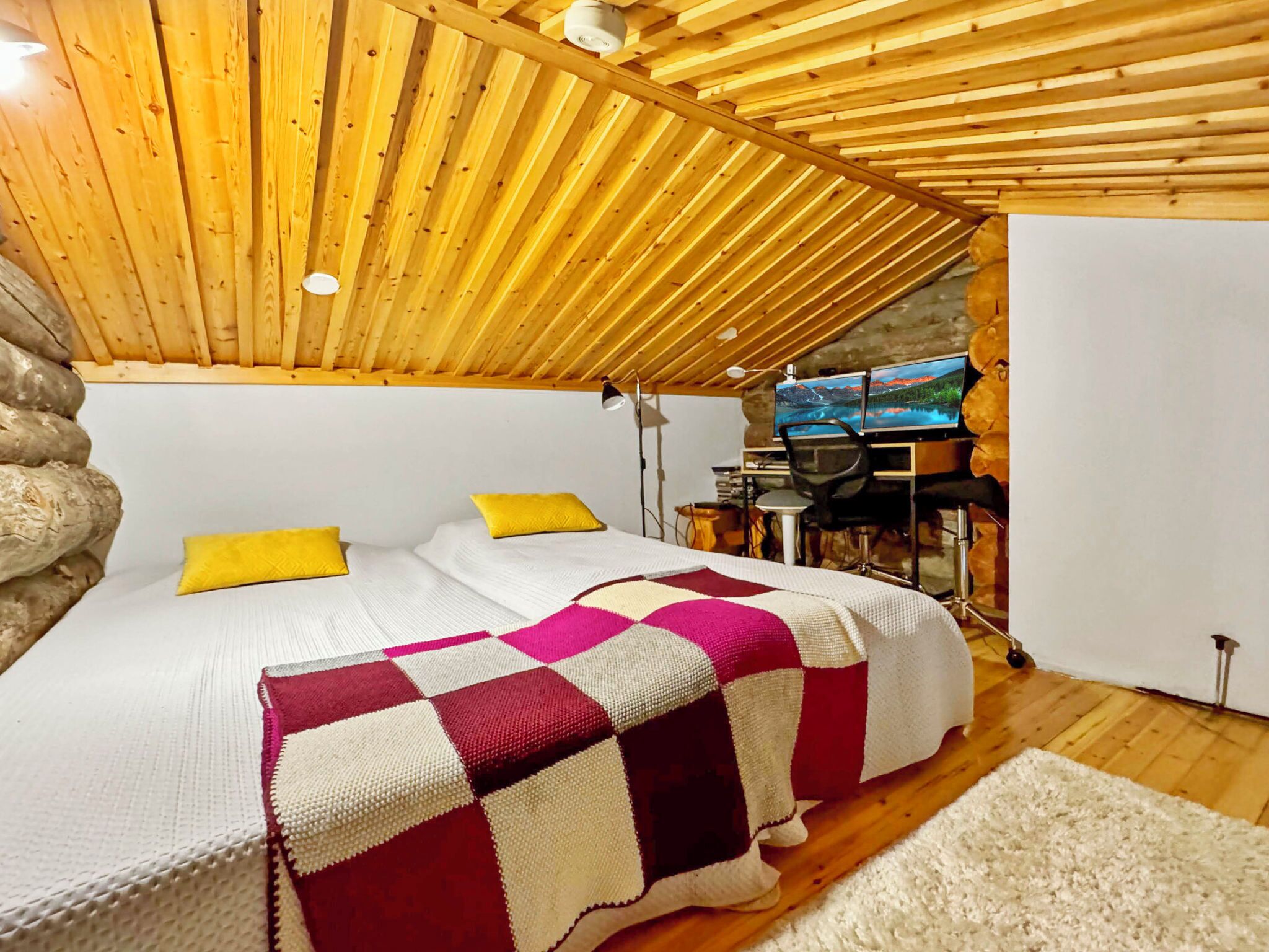 Een gezellige kamer met een houten plafond, een bed met een geruit plaid, een bureau met een computer en een stenen muur.