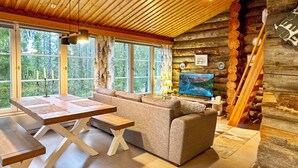 Interior de una cabaña de troncos con techo de madera, ventanas amplias, un sofá, una mesa de comedor y una televisión.