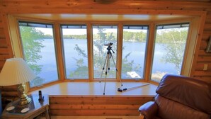 Interior de uma cabana de toras com uma grande janela em arco com vista para um lago, um tripé com uma câmera, uma poltrona de couro e um abajur sobre uma mesa de madeira.