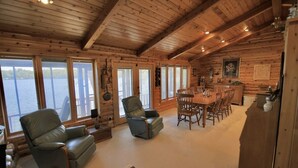 Interior de cabana de tronco com área de jantar, duas poltronas de couro reclináveis, cozinha e grandes janelas com vista para um corpo de água.
