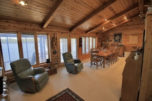 L’intérieur d’une cabane en rondins comprend un coin repas, deux fauteuils inclinables en cuir, une cuisine et de grandes fenêtres donnant sur un plan d’eau.