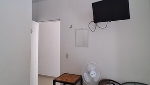 Una stanza con una TV a schermo piatto fissata al muro, una ventola a piantana, un tavolino di legno, una sedia con un cuscino colorato e una porta che dà su un'altra stanza.