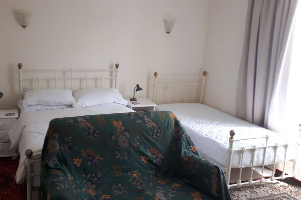 A single bed with a white headboard, a bedside table with a lamp, and a chair with a floral cover.