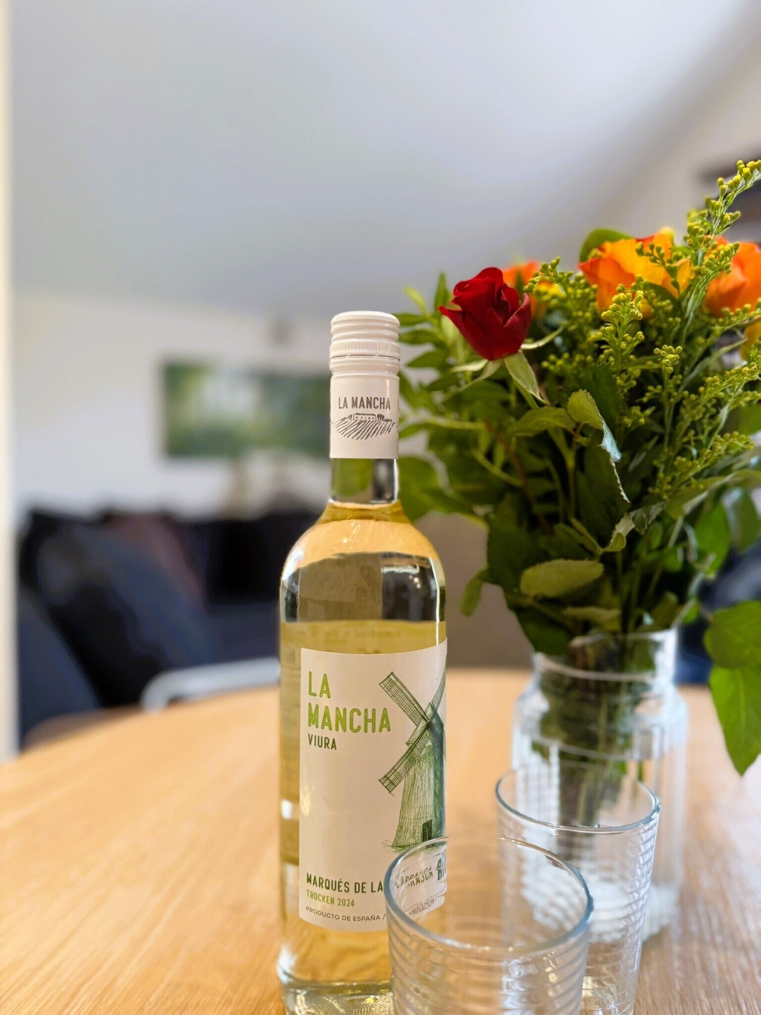 Pullo La Manchan viiniä, lasinen maljakko, jossa on kukkia, ja kaksi tyhjää lasia puisella pöydällä.