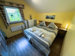 A bedroom with a bed, a window with curtains, a radiator, a bedside table with a lamp, and a painting on the wall. - Peaceful COZY Apartment in Bavarian Forest +WIFI (Grafenwiesen)