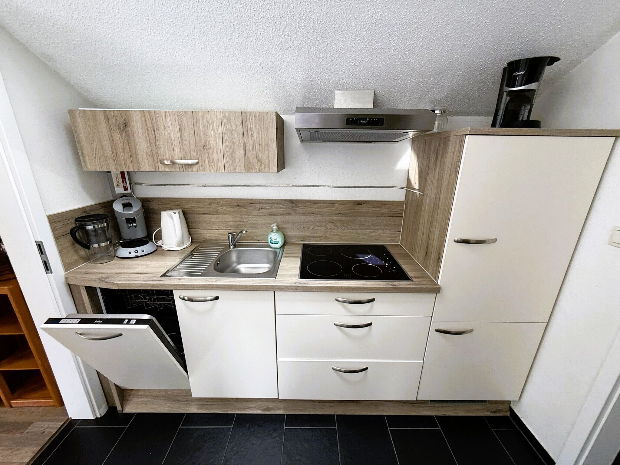 A compact kitchen with a built-in dishwasher, a coffee maker, a kettle, a sink, and a stove.