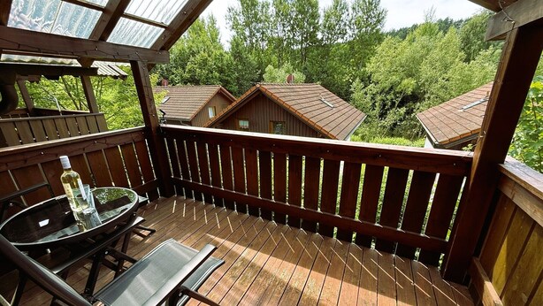 A wooden deck with a glass table and chairs, overlooking a forested area with houses. - Peaceful COZY Apartment in Bavarian Forest +WIFI (Grafenwiesen)