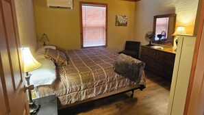 Ein Schlafzimmer mit Holzboden, einem Bett mit Steppdecke, einem Nachttisch mit Lampe, einer Kommode, einem Fenster mit Jalousien und einem Deckenventilator.
