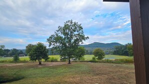 Blick von einer überdachten Fläche auf ein Wiesfeld, Bäume und ferne Hügel.