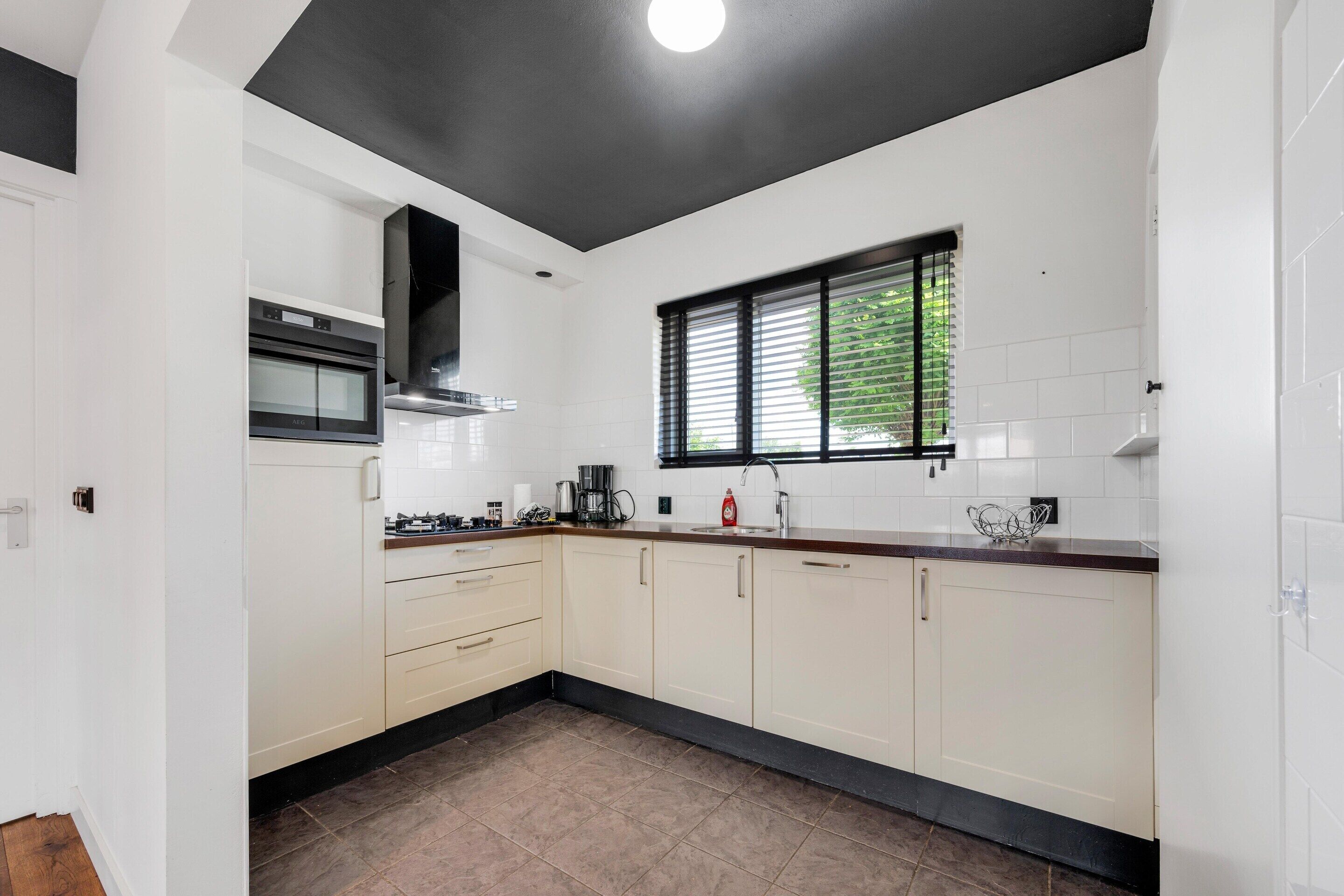 A modern kitchen with white cabinets, a black countertop, a window with blinds, and a stainless steel range hood.