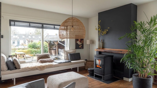 Ein modernes Wohnzimmer mit einer Couch, einem Hocker, Sesseln, einem Kamin, einem großen Fenster und einer Zimmerpflanze.