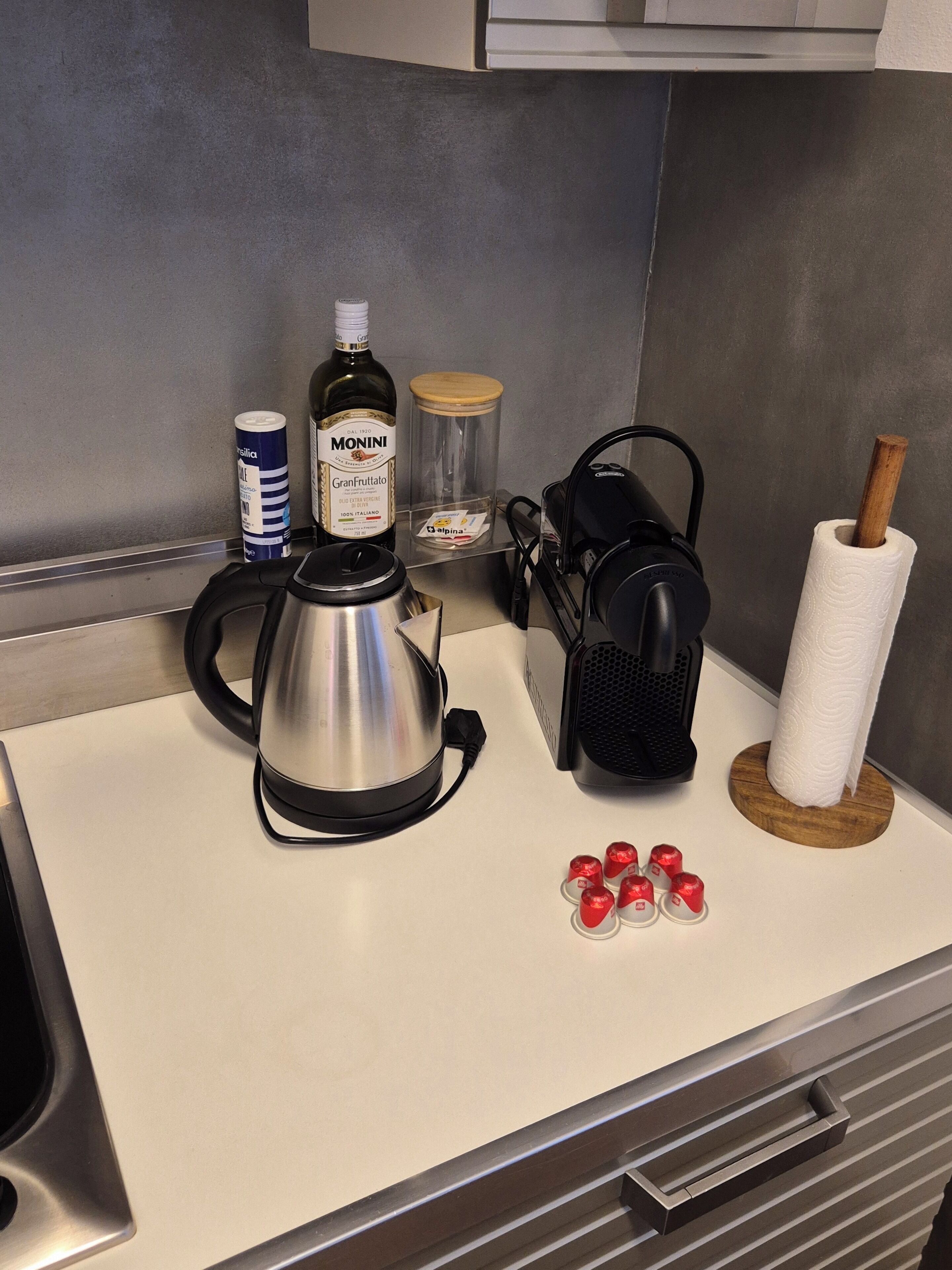 Eine Küchenarbeitsplatte mit einer Kaffeemaschine, einer Flasche Monini GranFruitatto, einer Dose Bisazza, einem Behälter Alpina und einer Rolle Küchenpapier.