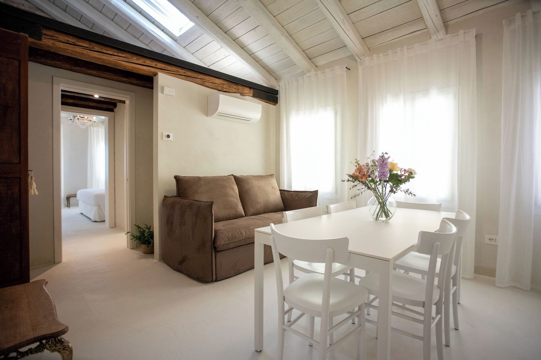 Una habitación con una mesa blanca, cuatro sillas, un sofá marrón y un florero con flores.