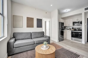 A modern living room with a grey sofa, a wooden coffee table, and a kitchen with stainless steel appliances.
