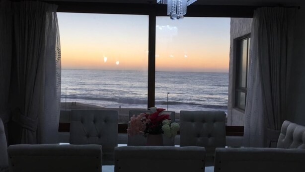 Een kamer met uitzicht op zee bij zonsondergang, met een tafel met bloemen en vier gestoffeerde stoelen.