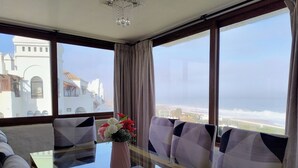 Een kamer met een glazen tafel, twee stoelen, een vaas met bloemen en uitzicht op gebouwen en de zee.