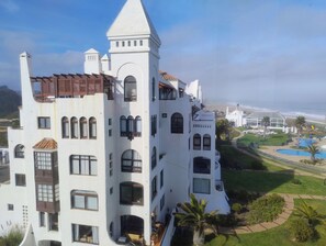 Un edificio grande y blanco de varios pisos con una torre destacada, numerosas ventanas y un césped cuidado con piscina.