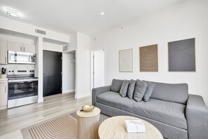 Ein modernes Wohnzimmer mit einem grauen Sofa, einem runden Holztisch und einer Küchenzeile mit Mikrowelle und Backofen.