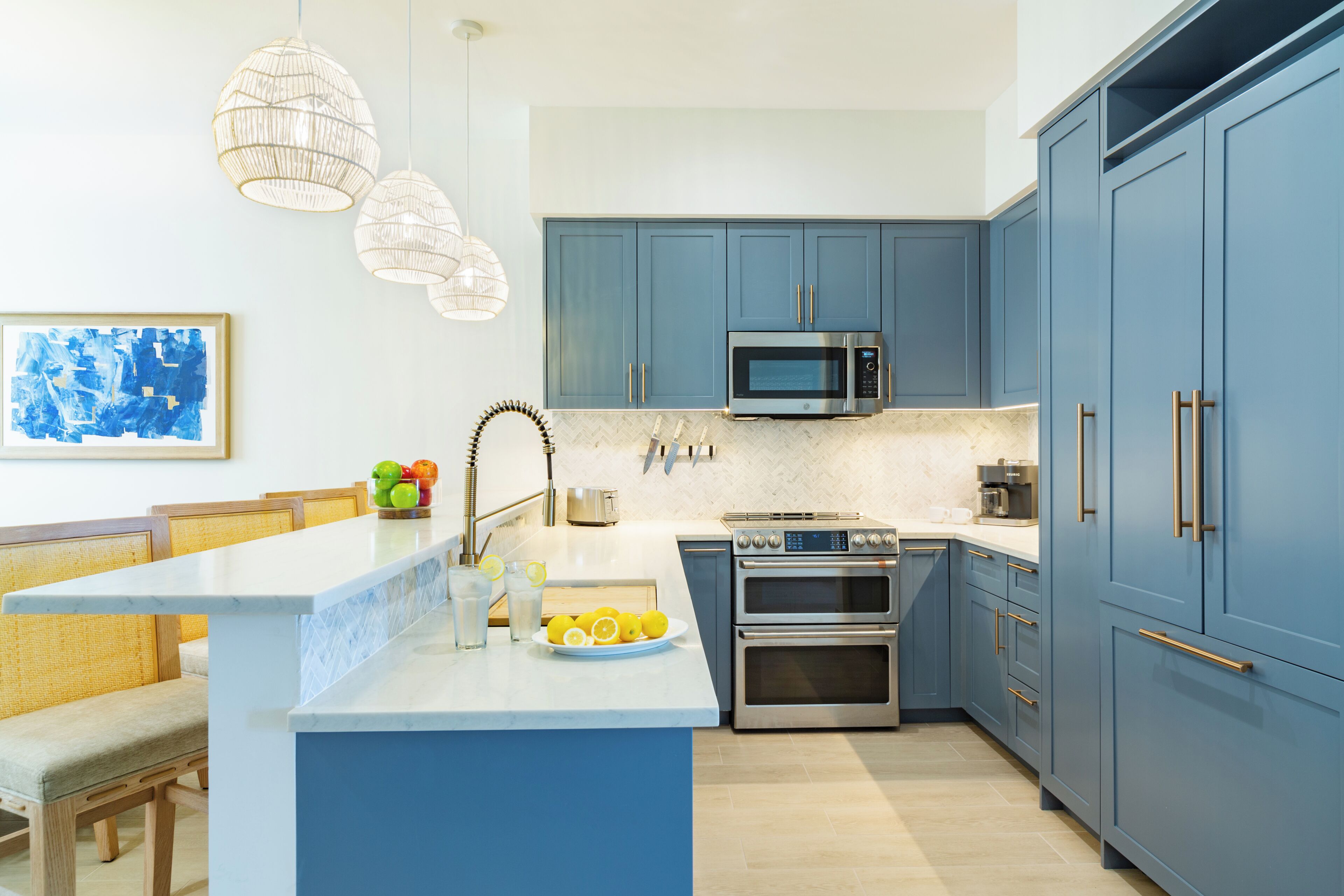 A modern kitchen with blue cabinets, a white countertop, a built-in oven, a microwave, and a dining area with a bench and table.