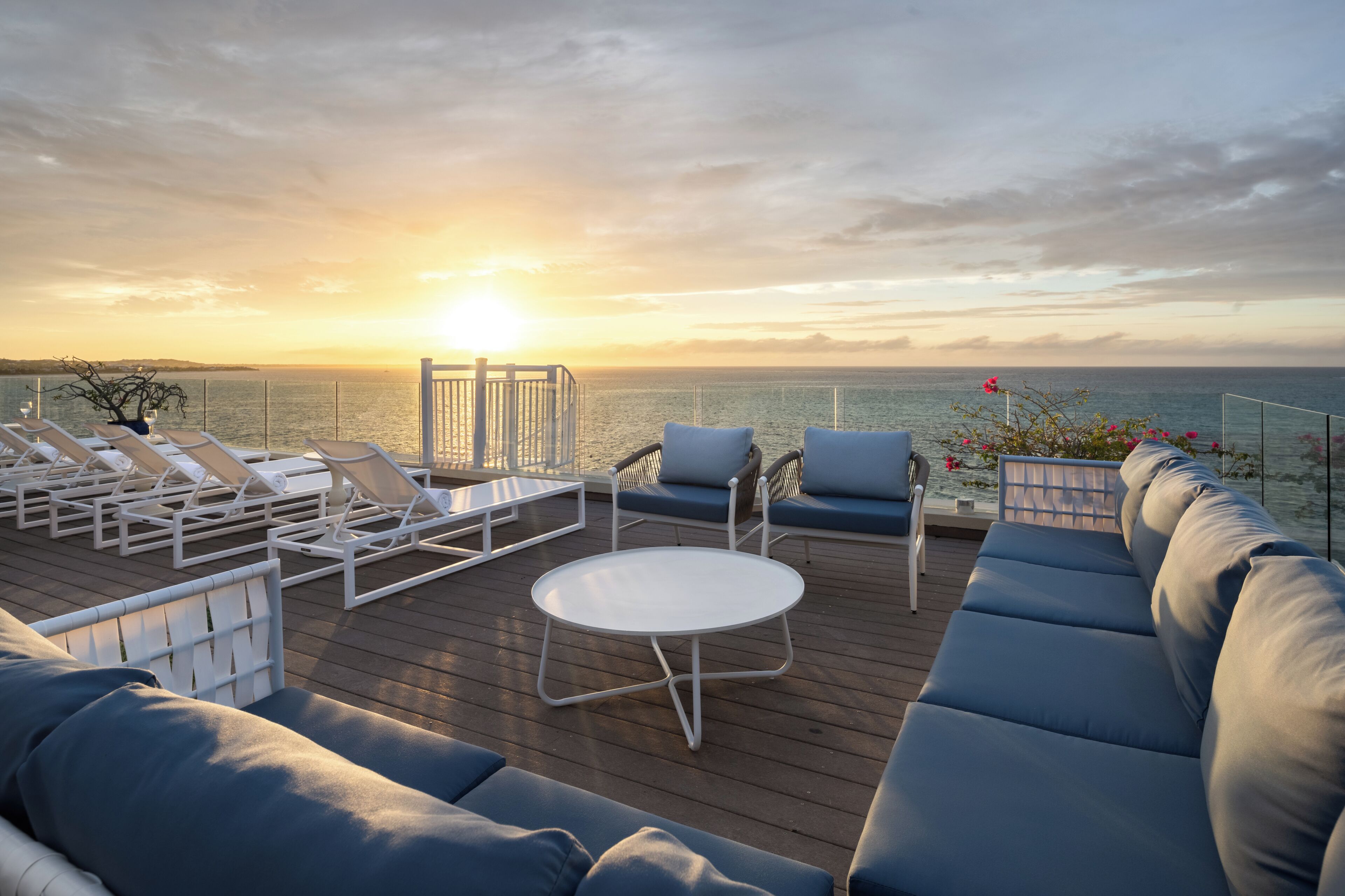 Een terras met loungebedden, een bank en een tafel, met uitzicht op zee tijdens de zonsondergang.