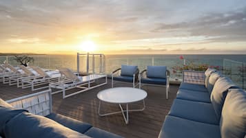 Een terras met loungebedden, een bank en een tafel, met uitzicht op zee tijdens de zonsondergang.