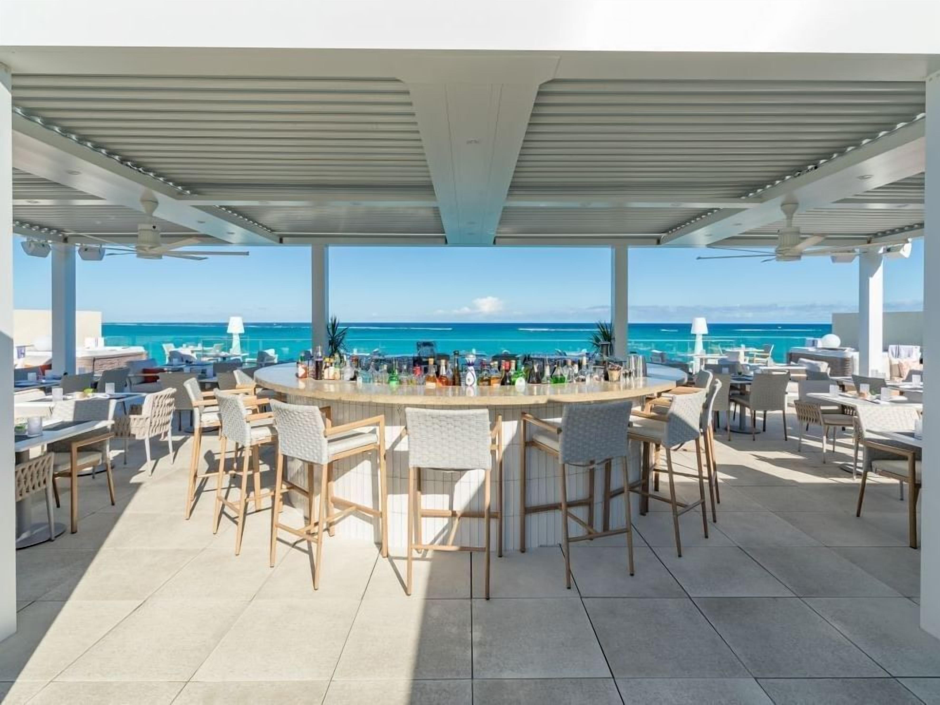 A covered outdoor bar area with a view of the ocean, featuring multiple seating arrangements and a central bar.