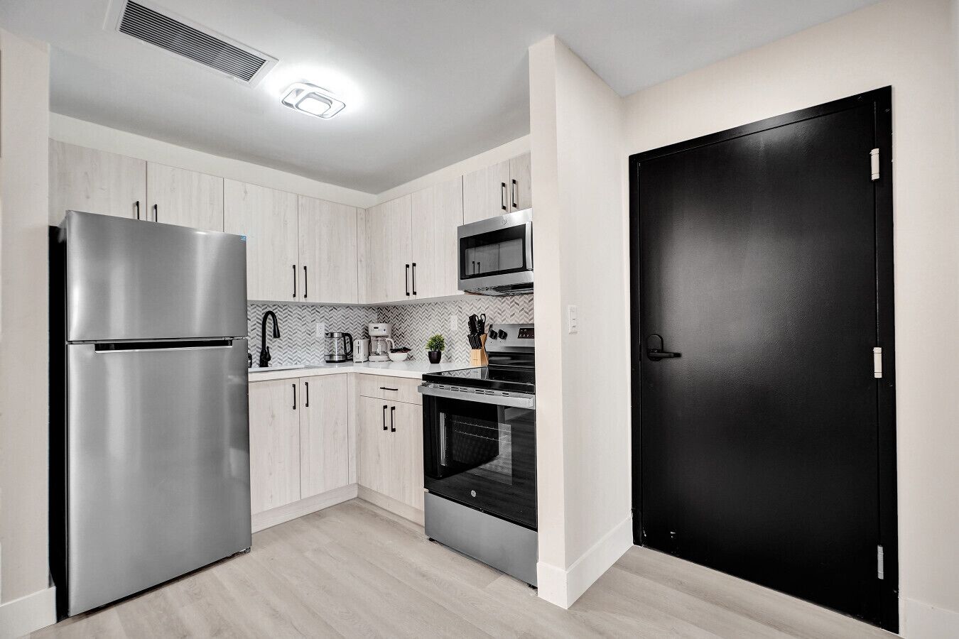 A modern kitchen with stainless steel appliances, light wood cabinets, and a black door.