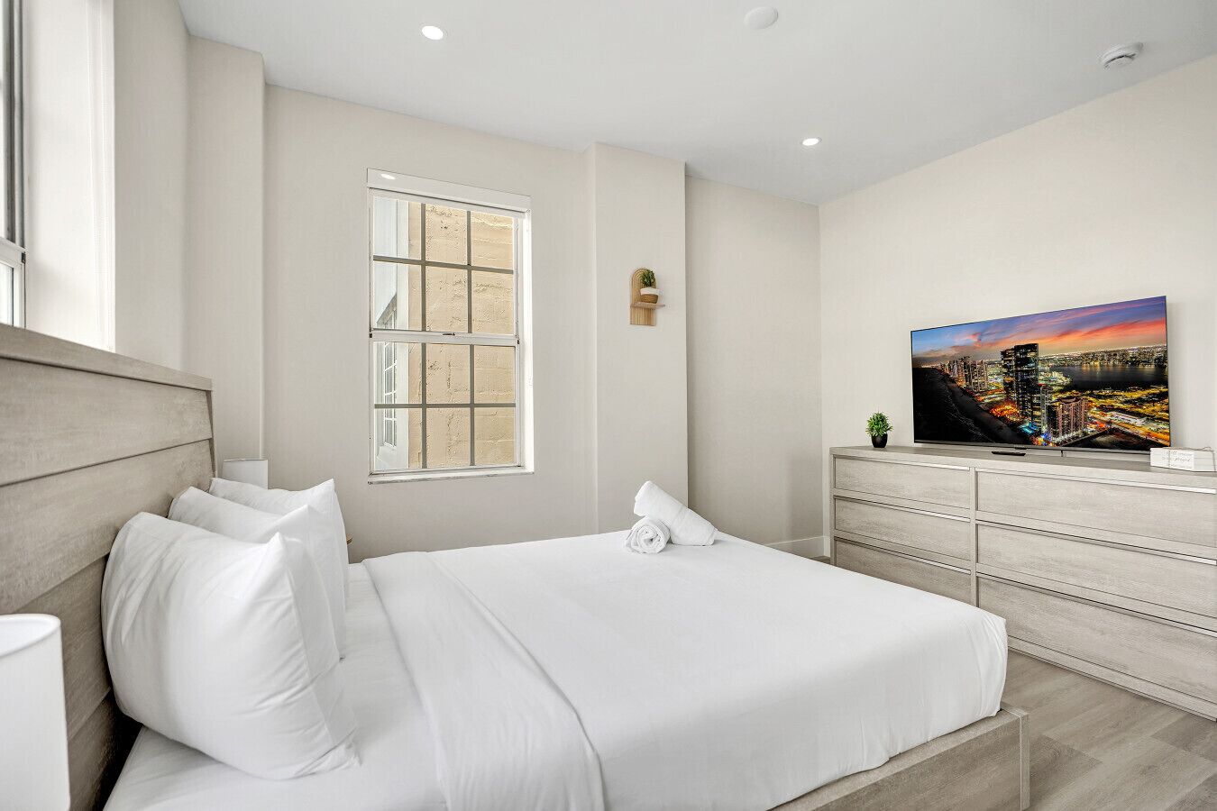 A modern hotel room with a large bed, a TV, a window with a city view, and a small plant on the dresser.