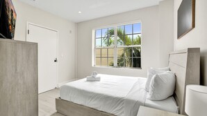 Una habitación de hotel moderna con una cama grande, una mesita de noche, una ventana con vistas a palmeras y una puerta.