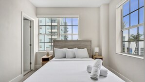 Une chambre d’hôtel avec un grand lit, deux tables de chevet, une fenêtre donnant sur des immeubles et une porte.