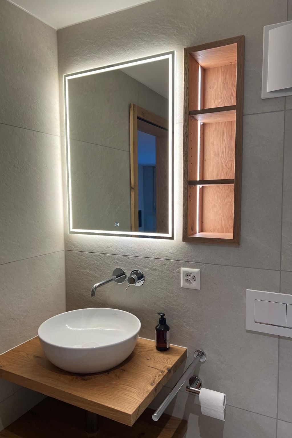 Un baño con un lavabo blanco, un espejo con iluminación LED, un armario de madera para medicinas y un perchero para toallas.