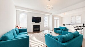 A modern living room with teal sofas, a glass coffee table, a fireplace, a flat-screen TV, and a chandelier. - Min 30 days Entire large 4bd house with 3 parking  (East Gwillimbury)