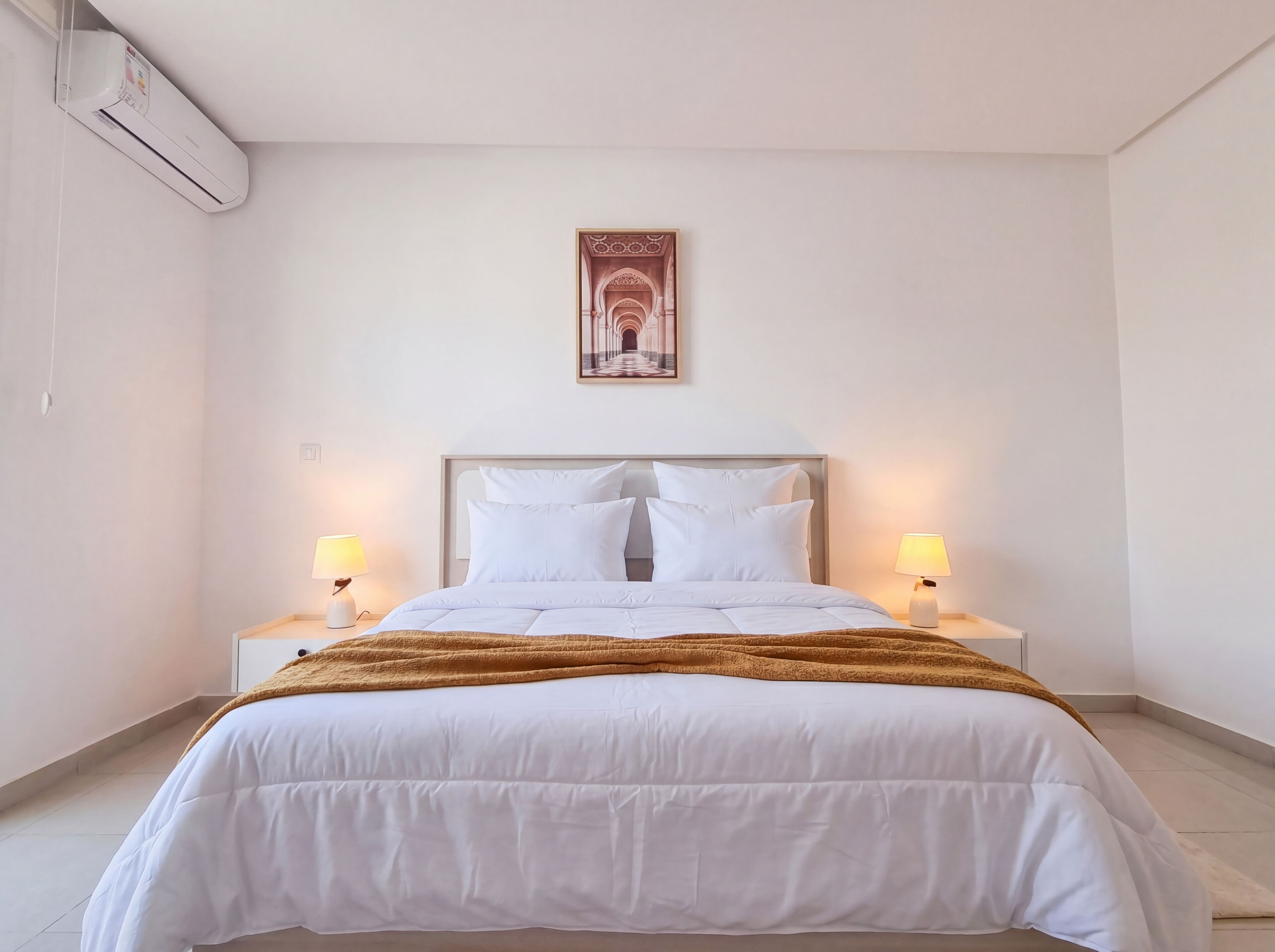 A neatly made bed with white bedding and a brown throw blanket, two bedside lamps, and a framed picture on the wall.