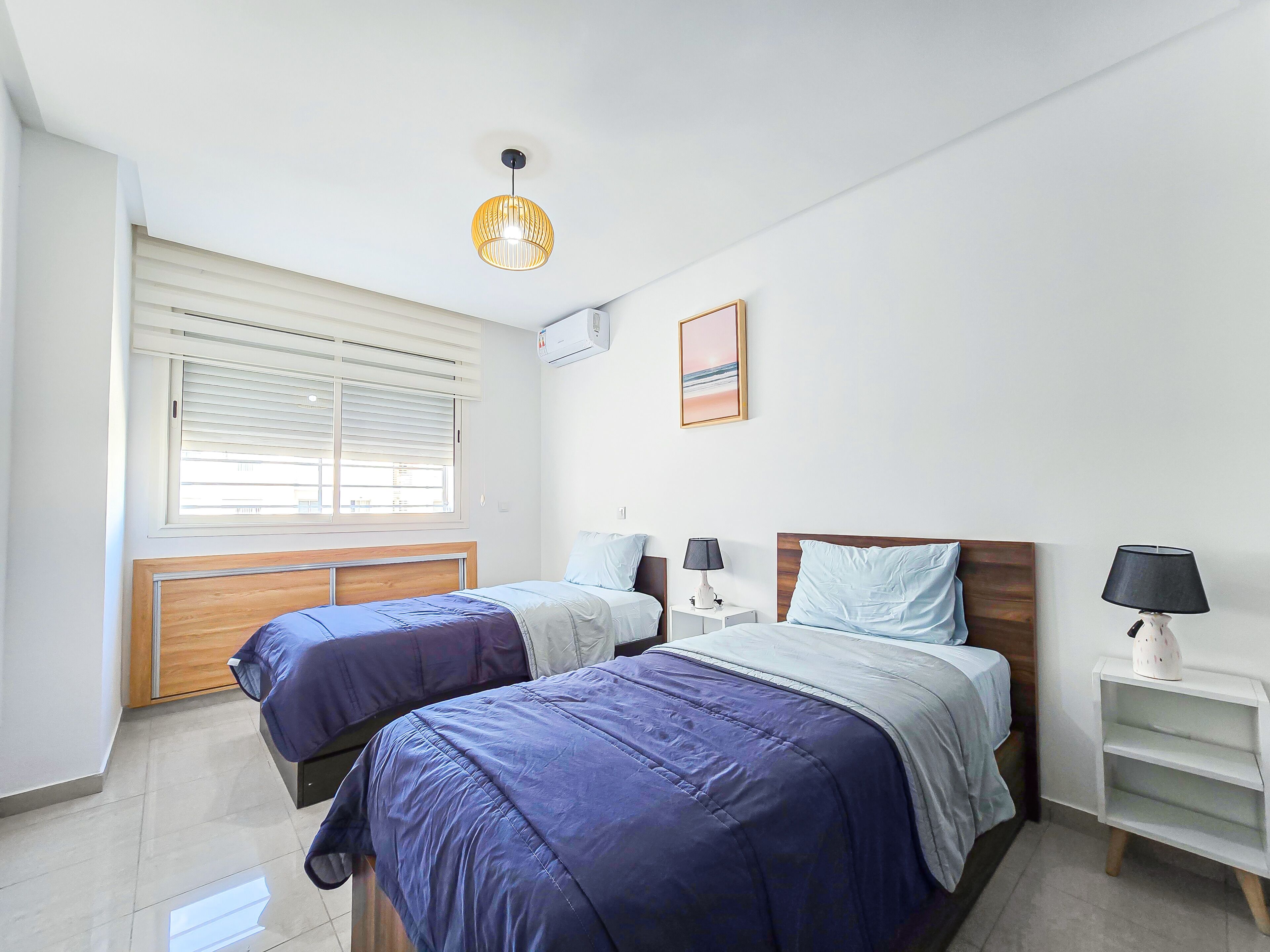 A bedroom with two beds, a window with blinds, a ceiling light, a wall-mounted air conditioner, and a bedside table with a lamp.