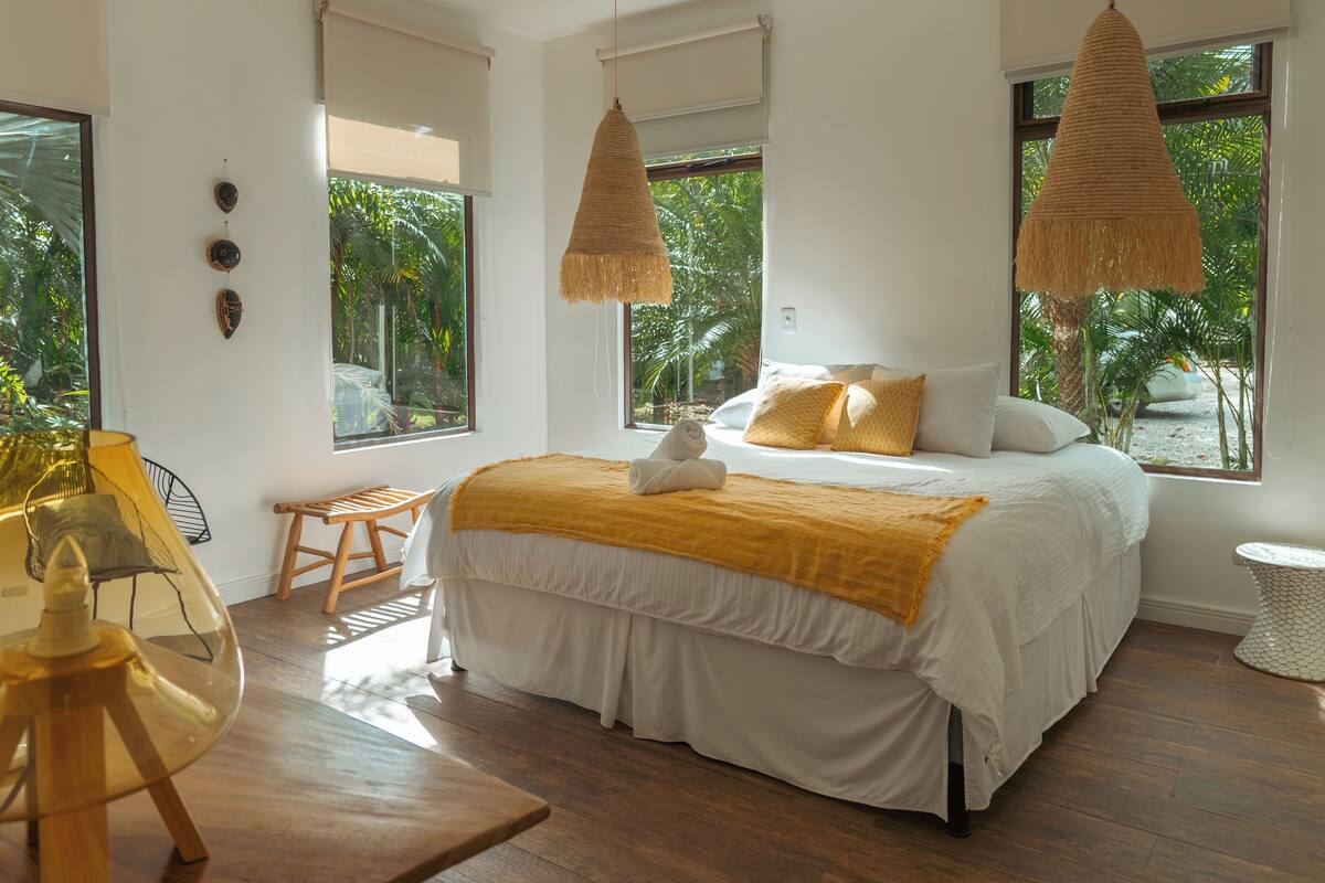 A bedroom with a bed, a hanging light, a chair, and a window with a view of greenery.