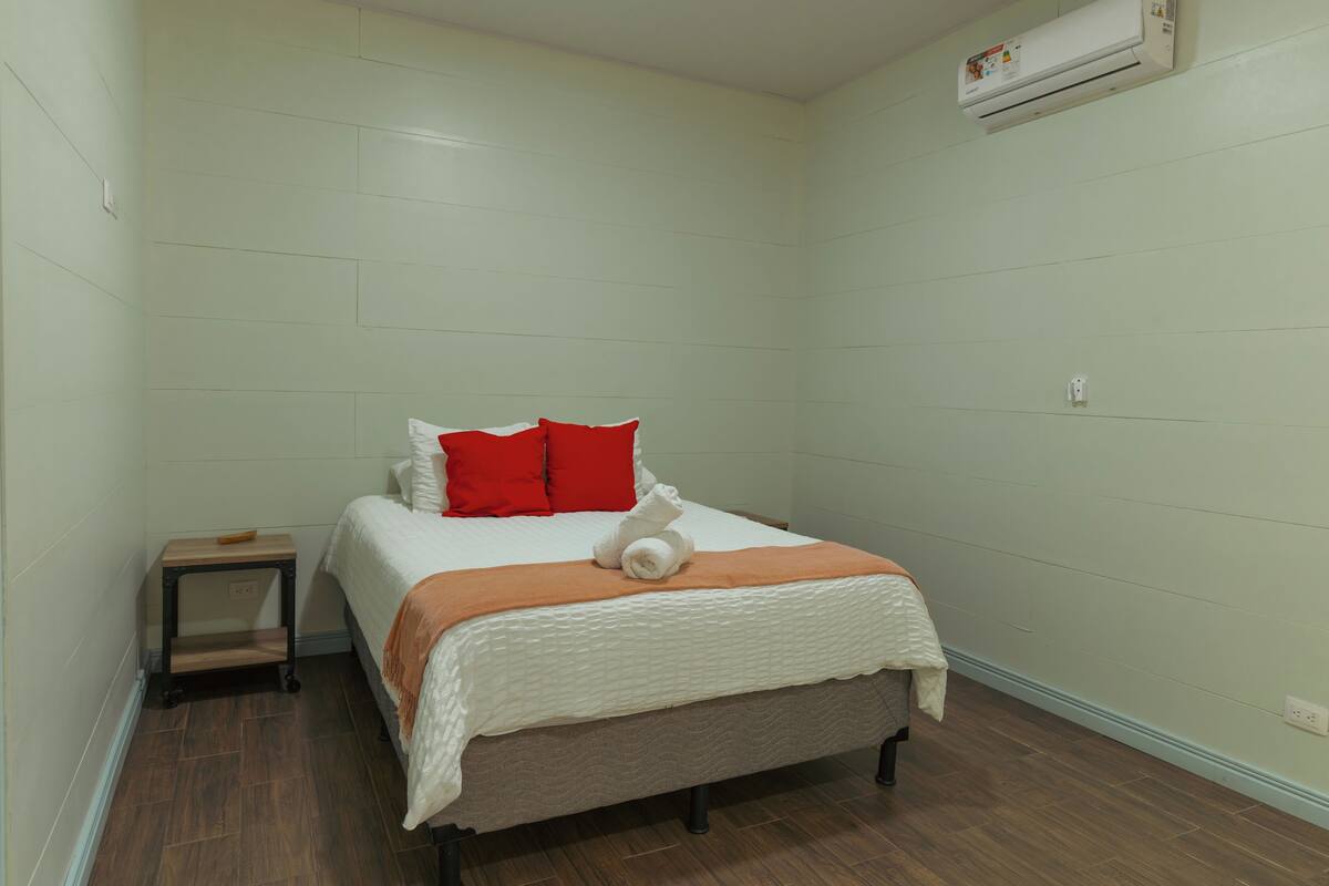 A small, neatly arranged bedroom with a bed, pillows, a bedside table, and an air conditioning unit.