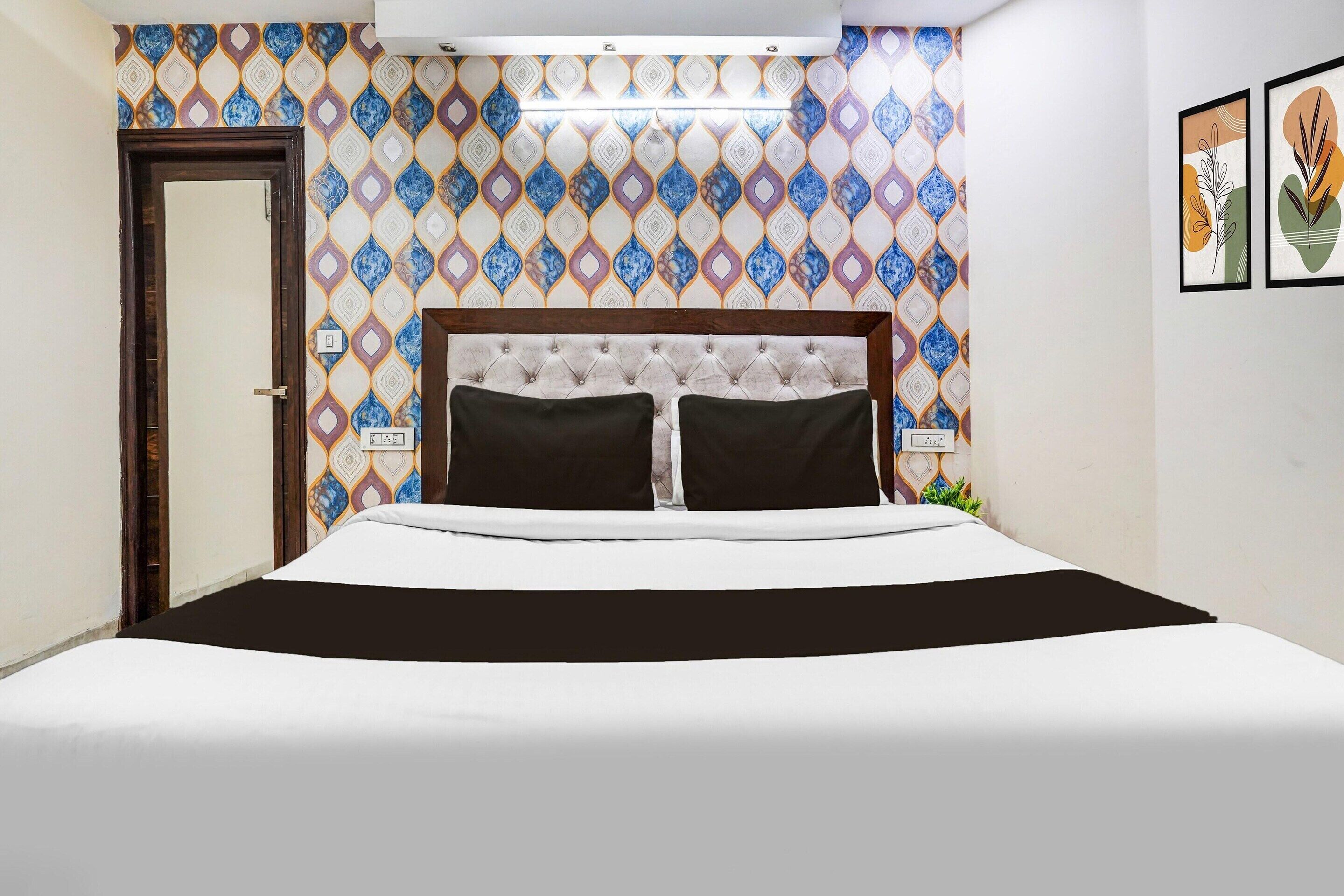 A hotel room with a patterned wall, a bed with a tufted headboard, two black pillows, and a bedside table with a lamp.