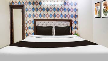 A hotel room with a patterned wall, a bed with a tufted headboard, two black pillows, and a bedside table with a lamp.
