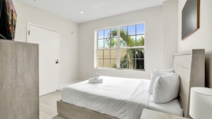 A modern hotel room with a large bed, a bedside table, a window with a view of palm trees, and a door. - Chic 1BR in the Heart of SoBe Ocean 1 Min Away 308 (Miami Beach)